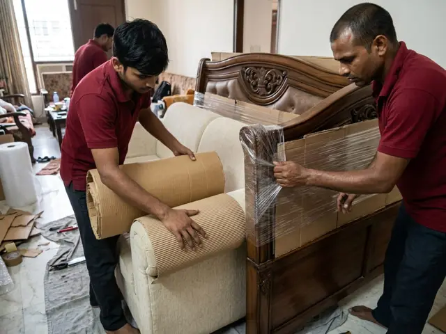Two packers wrapping a wooden bed headboard and sofa in stretch film and corrugated sheets